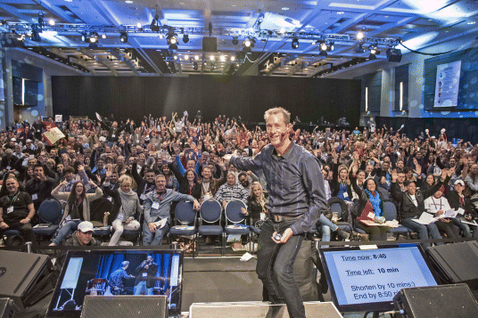 A photograph of David Meerman Scott addressing a huge crowd in high energy. He is pointing his right arm toward the audience and has a disc-shaped object in his left hand. Maximum audience are enjoying with a smile on their faces and raising their arms in the air.