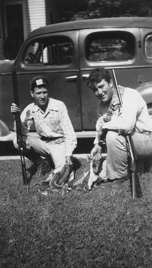 A photograph depicting Booker (right) and his friend in a crouching position displaying their hunt while holing their rifles. Behind them is a car.